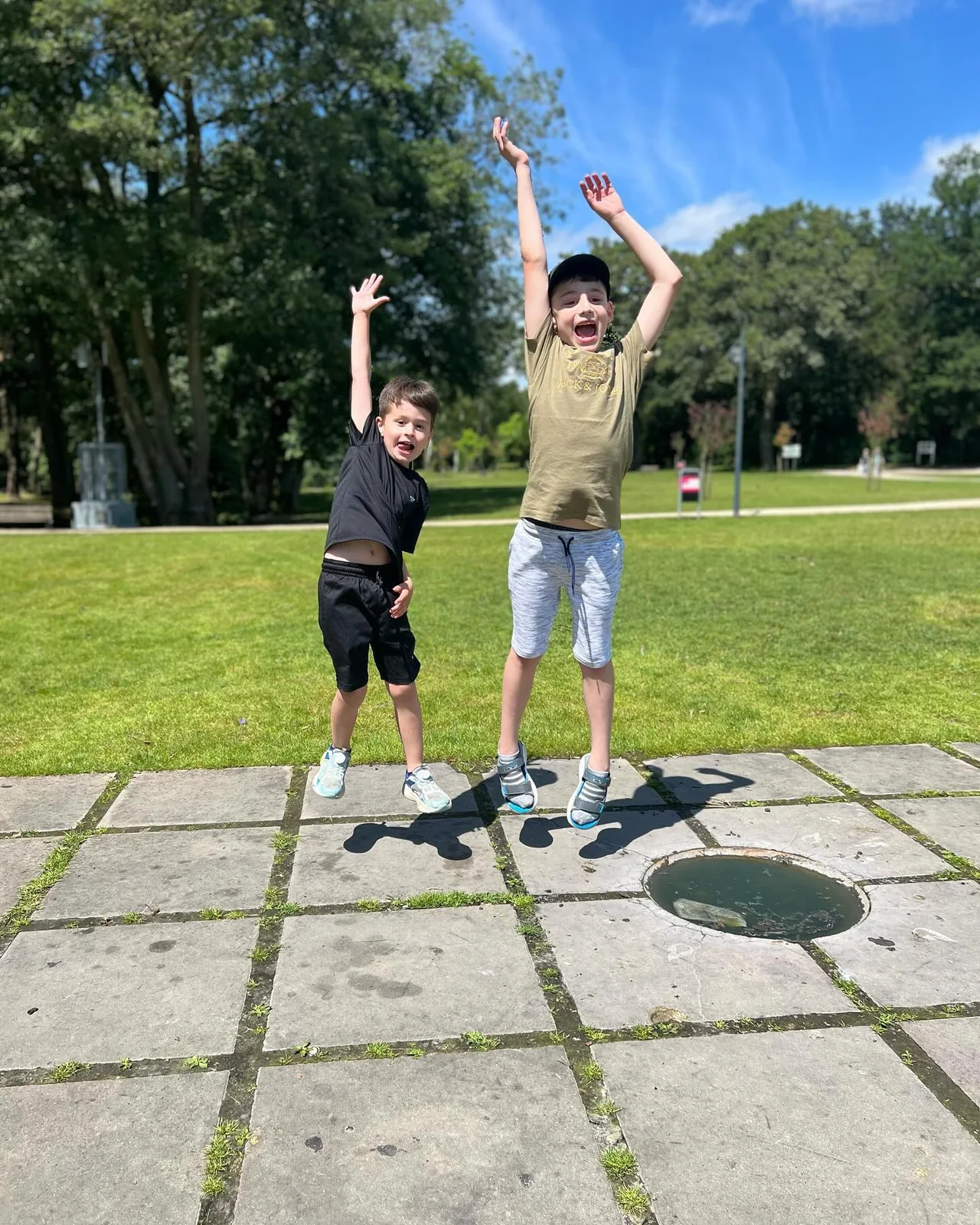 Twee deelnemers springen met hun armen omhoog op een grasveld naast een betegeld pad.