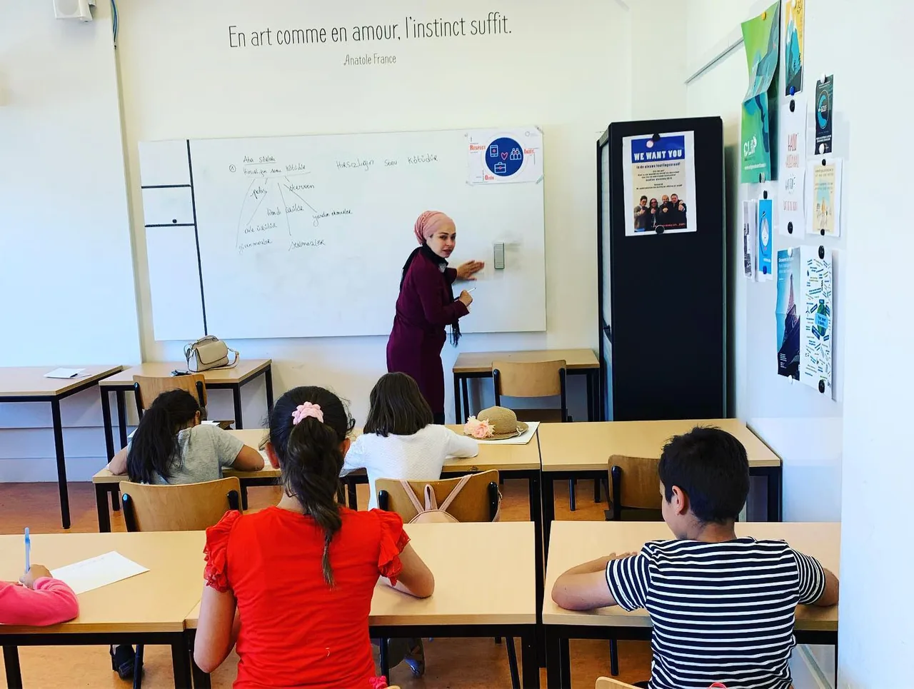 Kinderen zitten in een klaslokaal terwijl een begeleider uitleg geeft bij een whiteboard.